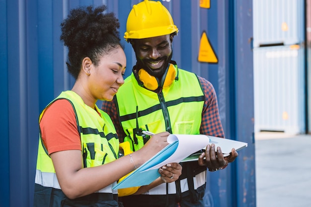 Сотрудник таможенной службы афроамериканской пары, работающий вместе с контрольным списком заказов на доставку на контейнерной площадке порта, наслаждается работой, счастливая улыбка