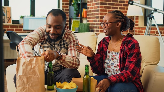 African american couple eating burgers with fries at home, having fun watching movie on tv and drinking beer. Boyfriend and girlfriend serving fast food meal from takeaway restaurant.