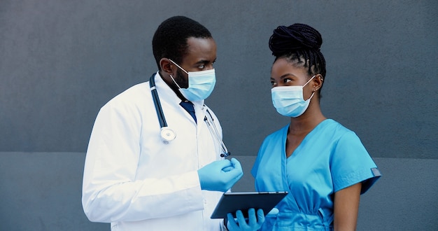 Coppia afroamericana dell'uomo e della donna, colleghi medici in maschere mediche lavorando e utilizzando il dispositivo tablet. medici maschii e femmine che parlano, toccano e scorrono sul computer del gadget.