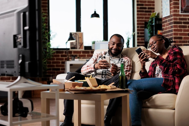 African american couple using smartphones and eating takeaway
food while they watch movie on television. takeout pizza, noodles,
burger and snacks with bottles of beer watching tv film.