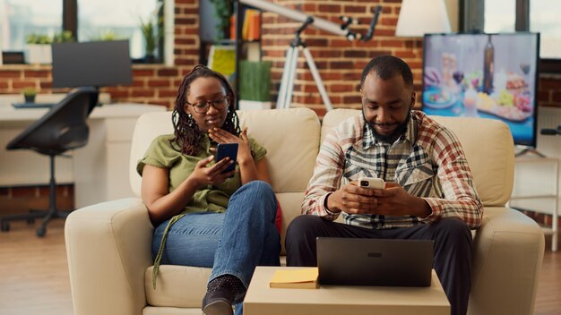Афроамериканская пара вместе использует смартфоны на диване, просматривает Интернет и просматривает социальные сети. Влюбленные люди смеются над цифровыми гаджетами, наслаждаясь отдыхом, расслабляясь.
