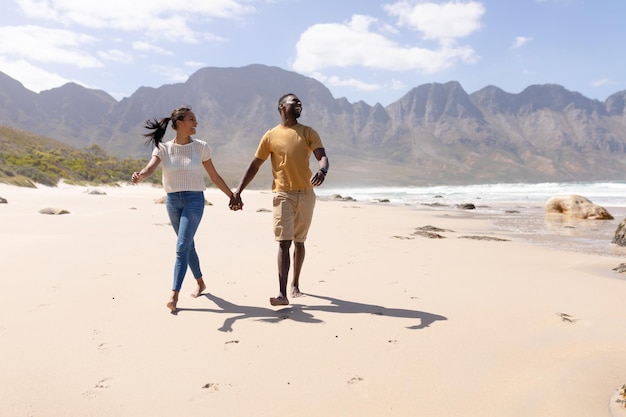 アフリカ系アメリカ人のカップルが海沿いのビーチで手をつないで歩いています。健康的なライフスタイル、自然の中でのレジャー。