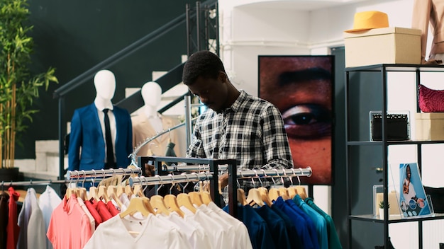 African american customer checking basic shirt, analyzing fashionable clothes fabric in modern boutique. Shopaholic man shopping for casual wear, looking at hangers with new fashion collection