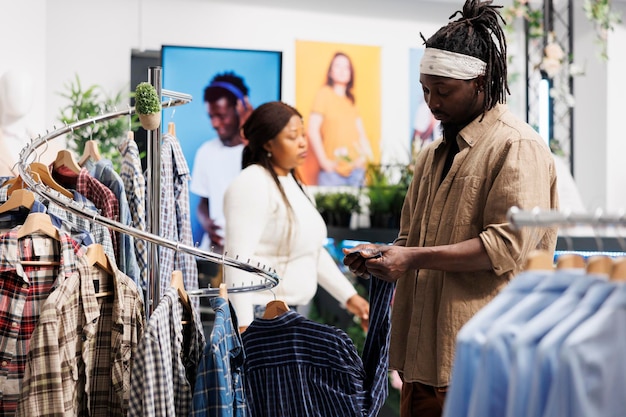 衣料品モールで購入する前にチェック柄のシャツの品質を検査するアフリカ系アメリカ人の顧客。ファッション ショールームの男性クライアントがフォーマルなアパレルを選択し、袖のラベルを探しています