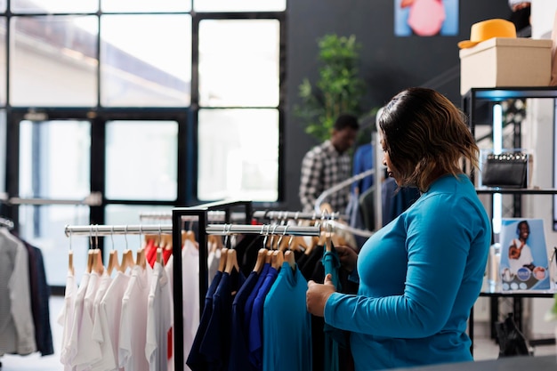 African american customer looking at hangers full with trendy clothes in modern boutique, shopping for trendy merchandise. Shopaholic woman checking new fashion collection, analyzing fabric