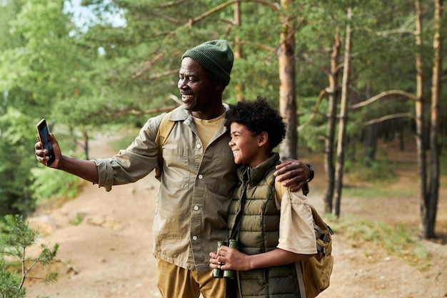 아프리카계 미국인 아빠와 아들이 숲에서 하이킹하는 동안 스마트폰으로 화상 통화를 합니다.