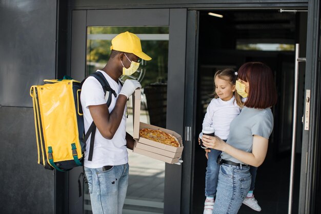 ピザと段ボール箱を提供する黄色のフェイス マスク制服キャップでアフリカ系アメリカ人の配達人