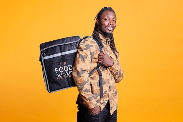 African american deliveryman carrying food delivery thermal backpack, standing in studio over yellow background. Restaurant courier deliverying healthy meal to customers during lunch time