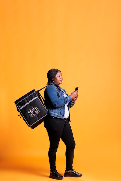 African american deliverywoman holding phone looking at customer adreess on fast food app before start deliver takeout order. Pizzeria courier carrying takeaway thermal backpack in studio