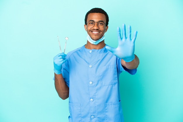 African American dentist holding tools over isolated blue background counting five with fingers