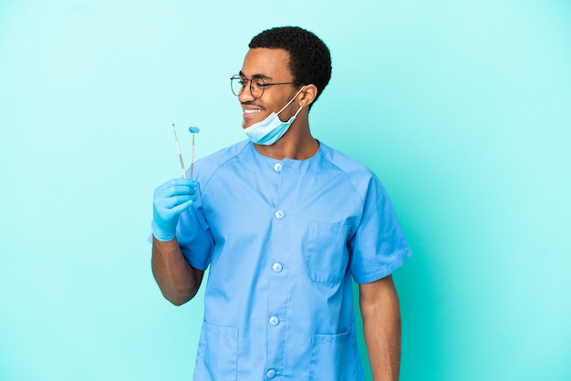 African American dentist holding tools over isolated blue background looking to the side and smiling