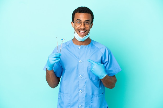 Dentista afroamericano che tiene gli strumenti su sfondo blu isolato con espressione facciale a sorpresa