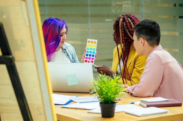 다양한 색상의 분홍색 파란색 긴 머리 소녀와 함께 앉아 있는 아프리카 계 미국인 디자이너는 회의실에서 프로젝트에 대한 새로운 아이디어를 제공합니다.