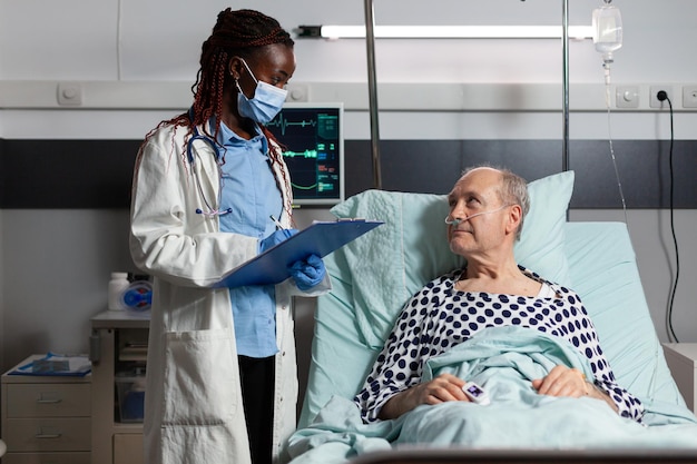 African american doctor with surgical mask reading diagnosis from clipboard to sick ill unwell senior patient laying in bed, breathing with help from oxygen mask, listening discussing with medical sta