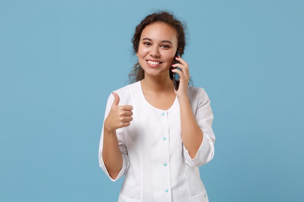 African american doctor woman isolated on blue background.\
female doctor in white medical gown talk on mobile phone showing\
thumb up. healthcare personnel health medicine concept. mock up\
copy space.