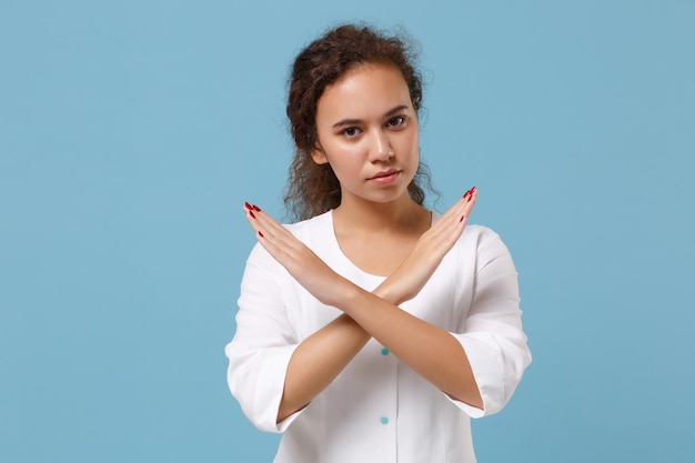 African american doctor woman isolated on blue wall background.\
female doctor in white medical gown showing stop with crossed\
hands. healthcare personnel health medicine concept. mock up copy\
space.