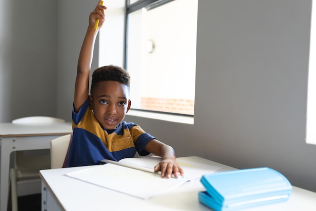 African american elementary schoolboy with hand raised sitting at desk in classroom