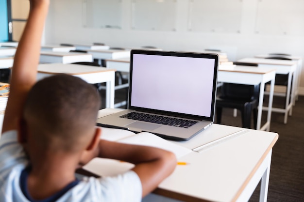 African american elementary schoolboy with hand raised using laptop at desk in classroom