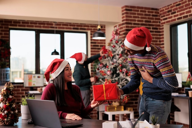 African american employee giving present to colleague, celebrating holiday season and christmas eve in festive office with seasonal decorations and ornaments. People feeling happy about xmas time.