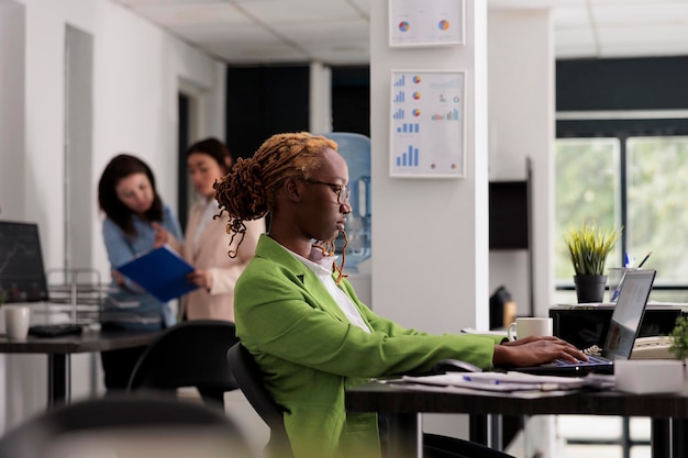 African american employee working on laptop in coworking space, manager sitting at workplace desk, colleagues on background. Young woman typing on computer, side view medium shot