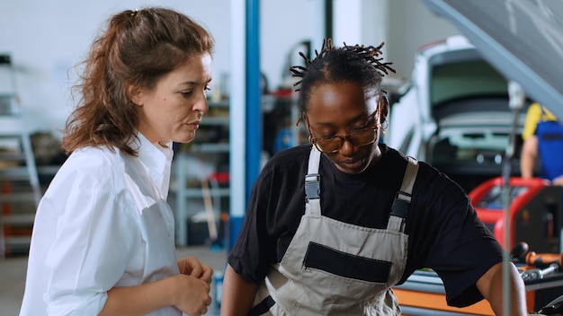 African american engineer assisting customer with car maintenance in repair shop Trained professional in garage looking over car components with woman servicing her vehicle during annual checkup