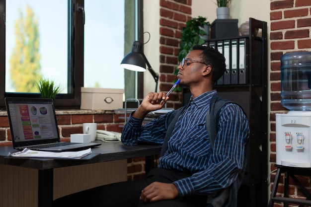 African american entrepreneur touching chin with pen looking focused at laptop screen with business analyitics. Startup employee thinking about sales results sitting at desk.