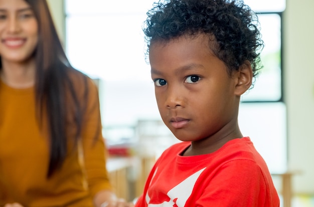 African American ethnicity kid with teacher smiling at background in kindergarten classroom