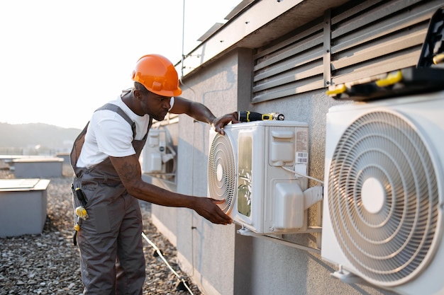 african american factory worker in orange hard hat standing and holding hands on air conditioner while checking attachment to wall outdoors concept of manual work and installation of equipment