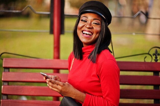 African american fashion girl in newsboy cap with handbag posed at street sitting on bench with mobile phone