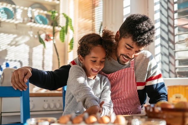 African american father and his daughter sitting in the kitchen and reading a recipe. Dad is wearing an apron. They are both happy.