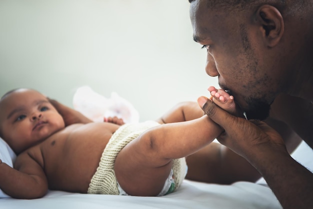 An African American father kissing foot of his baby newborn daughter is laying on the white bed