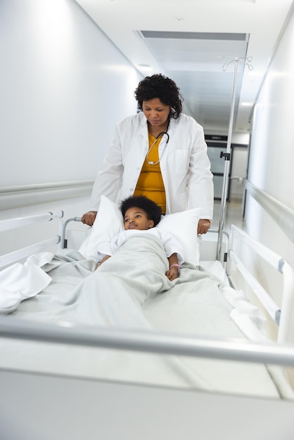 Photo african american female doctor walking with girl patient lying on bed in corridor at hospital. hospital, medicine and healthcare, unaltered.