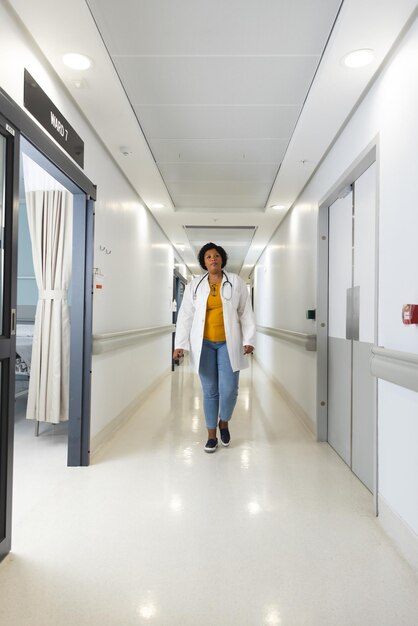 African american female doctor wearing lab coat and stethoscope walking in corridor at hospital. Hospital, work, medicine and healthcare, unaltered.