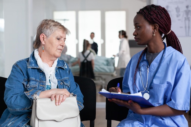 African american female doctor writing medication prescription for elderly sick woman in hospital lobby. Senior lady consulting general practitioner in geriatric clinic waiting area.