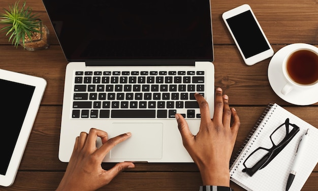 African-american female hands on laptop keyboard, blank screen for advertisement. Top view of black hands, coffee, smartphone, notebook on wooden table. Education, business, technology concept