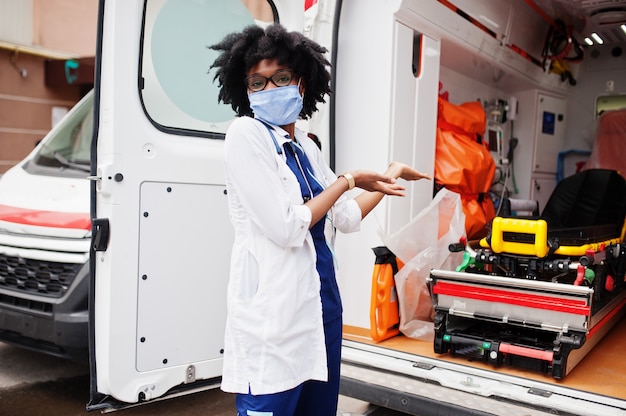 African American female paramedic in face mask standing in front of ambulance car