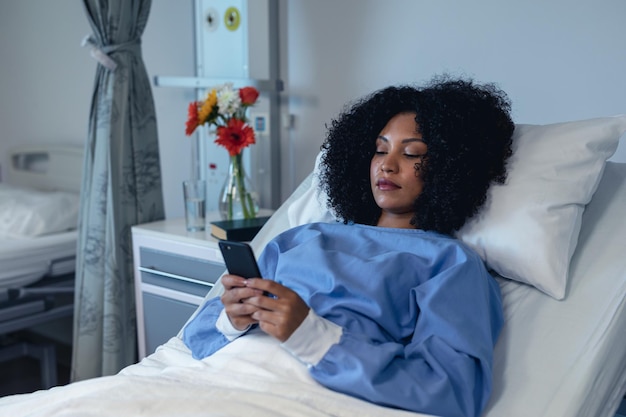 African american female patient lying in hospital bed using smartphone