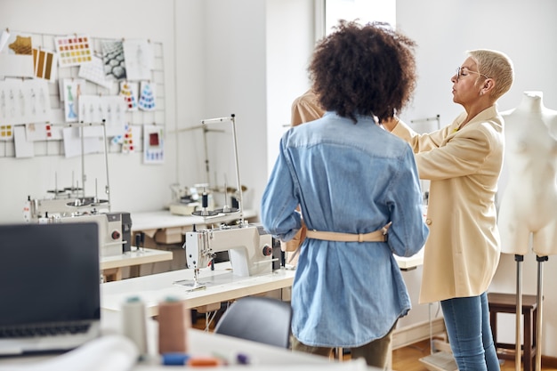 African-American female stagist looks at mature lady fashion designer working with new jacket on mannequin in light sewing workshop