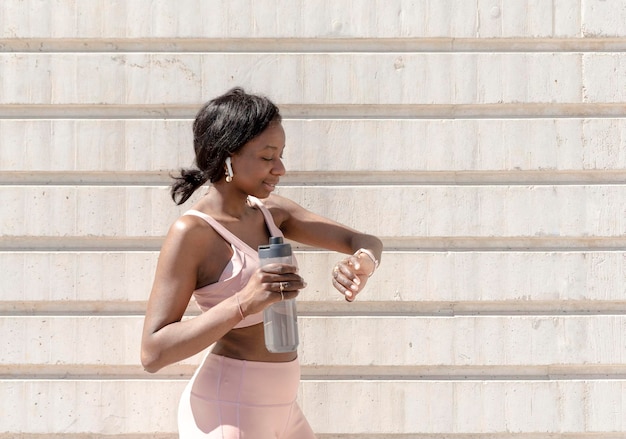 African American fit woman taking a break after running checking progress on smart watch and holding a bottle with water Runner looking at heart rate monitor on smart watch hydration