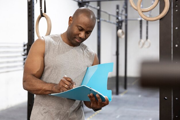 African American fitness instructor in gym. He is writing in a notebook. Space for text.