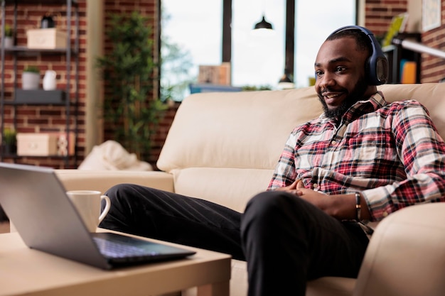 African american freelancer listening to music or podcast show\
on online website, using social media and headphones to work\
remotely. working on laptop at home and enjoying song on internet\
at home.