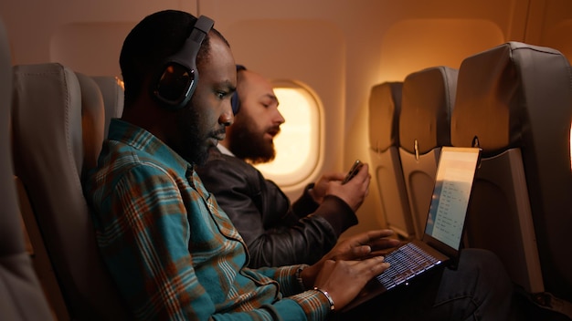 Libero professionista afroamericano che utilizza il laptop in volo, viaggiando in viaggio di lavoro durante il tramonto in classe economica. lavorare al computer e volare in aereo, viaggio aereo.