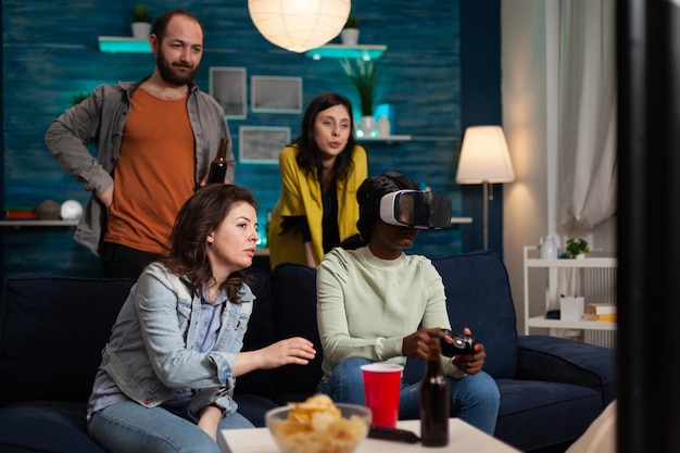 African american gamer woman wearing virtual reality headset playing videogames while friends guding her during online competition enjoying spending time together. Hanging out concept