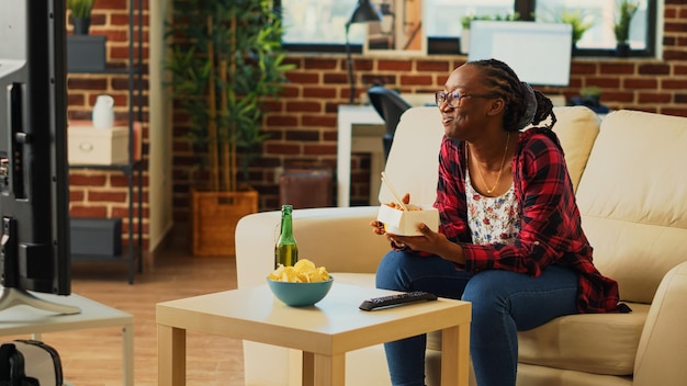 African american girl drinking beer and eating noodles in living room, having fun with delivery meal and film on television. Modern woman enjoying asian fast food and alcohol at home.