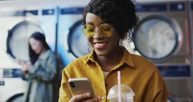 African american girl drinking orange juice with straw, texting\
message on phone and waiting for clothes to get clean woman sipping\
drink in laundry service room and tapping or scrolling on\
smartphone