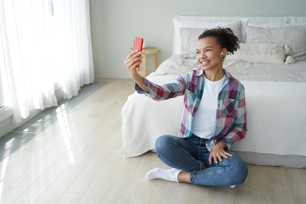 La ragazza afroamericana tiene il telefono in chat con un amico tramite videochiamata seduto sul pavimento in camera da letto