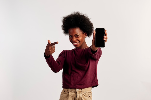 사진 갈색 스웨터를 입은 아프리카계 미국인 소녀가 스마트폰을 가리키며 포즈를 취하고 있습니다.