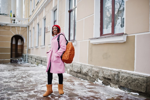 African american girl in red hat and pink coat with backpack at street of city on winter day.