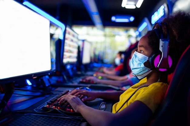 African American girl wearing face mask and headset playing video games on computer in game room