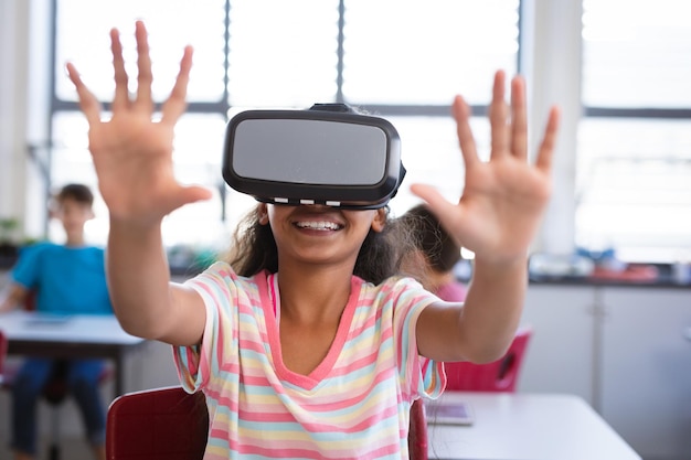 African american girl wearing vr headset gesturing while sitting on her desk in the class at school